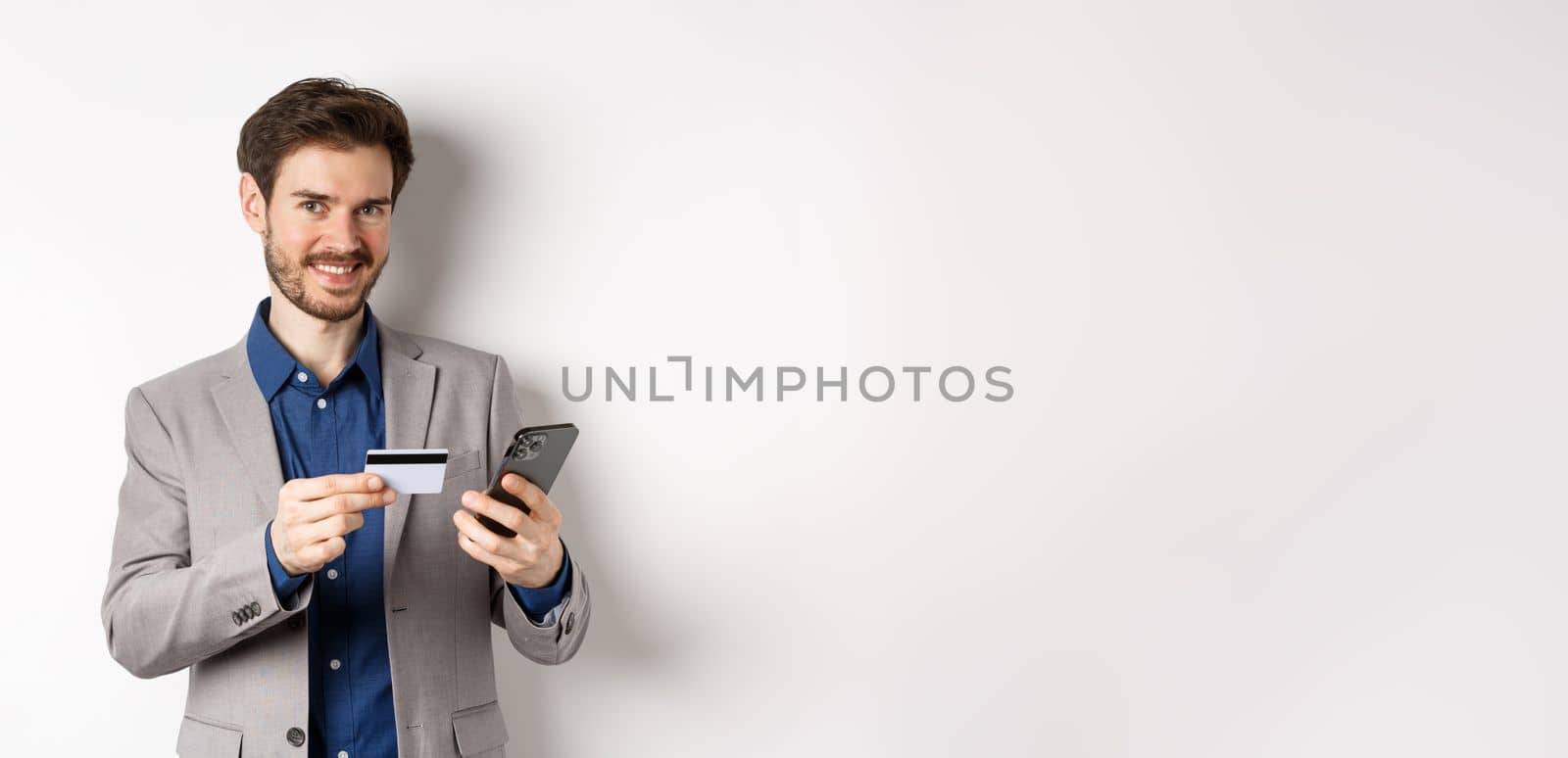 Online shopping. Handsome businessman in suit paying with credit card on smartphone, smiling satisfied at camera, standing on white background by Benzoix