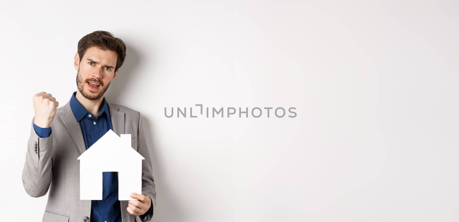 Real estate. Excited man buying house and showing paper home cutout, say yes and fist pump with joy, standing in suit on white background.