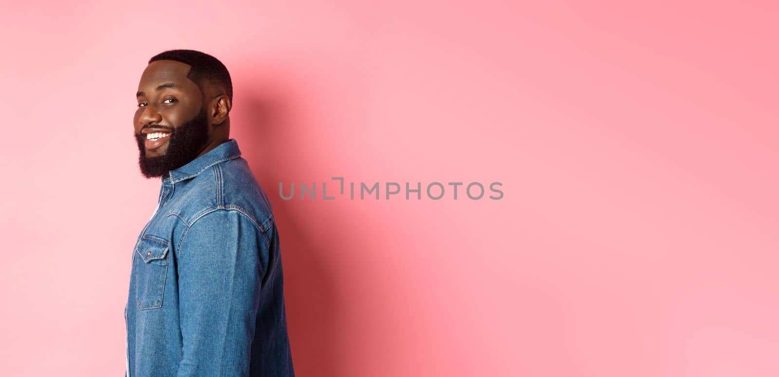 Handsome african-american man with beard, turn face at camera and smiling confident, standing over pink background.