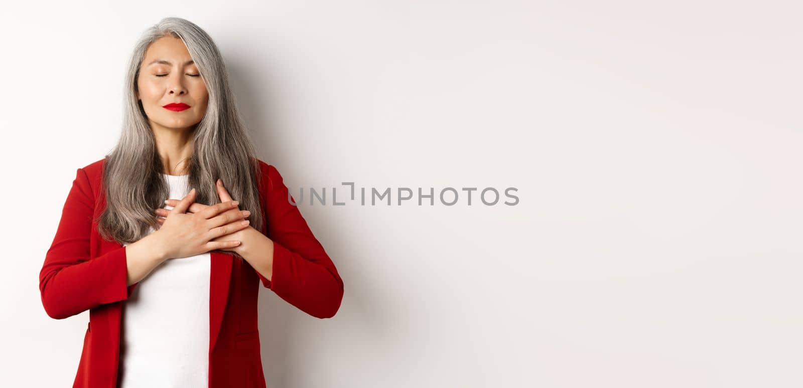 Senior asian woman with red lips and blazer, close eyes and holding hands on heart thankful, feeling nostalgic, dreaming of something, standing over white background.