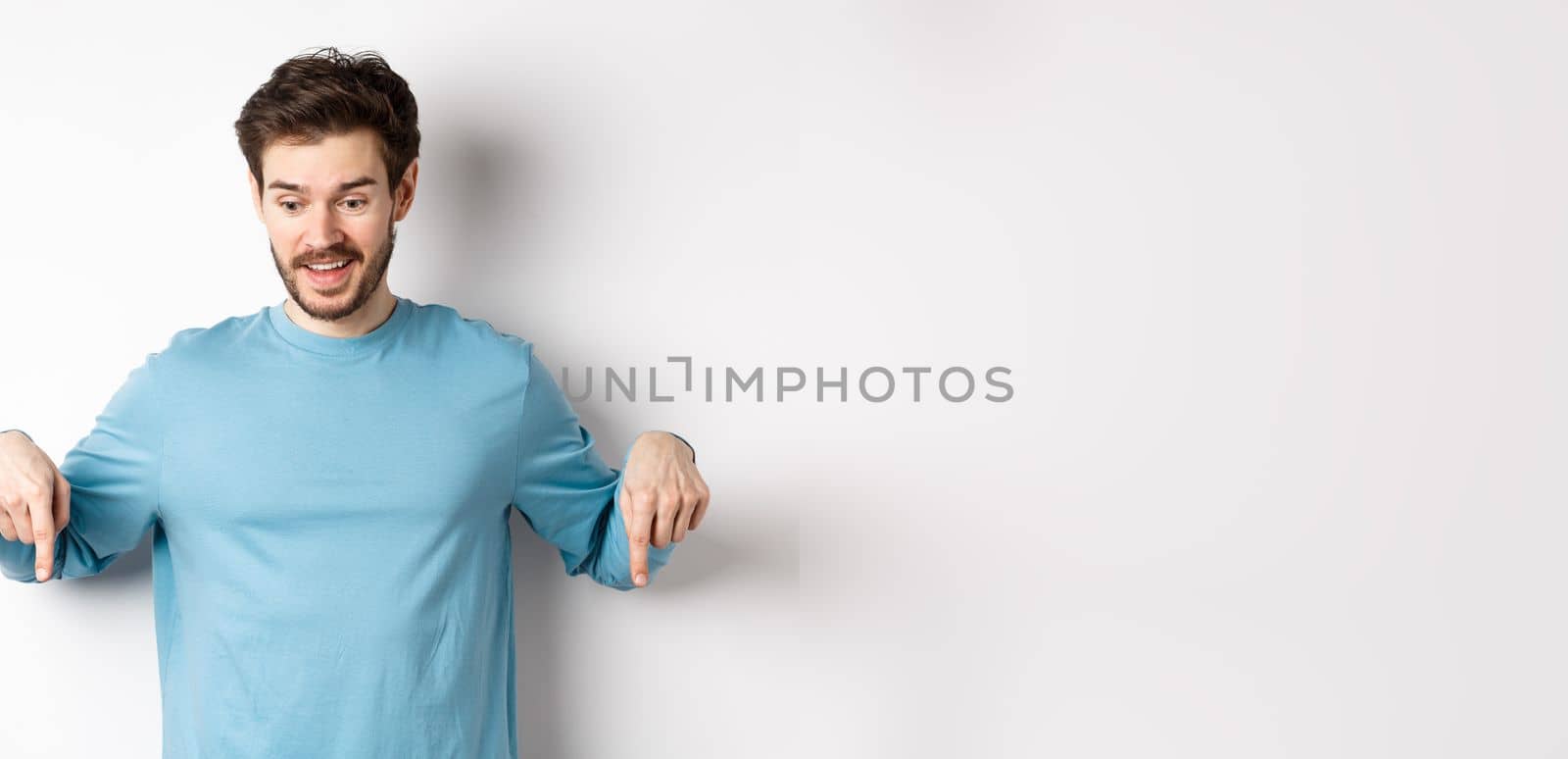 Excited and happy young man looking and pointing down at awesome deal, checking out promotion offer, standing on white background.