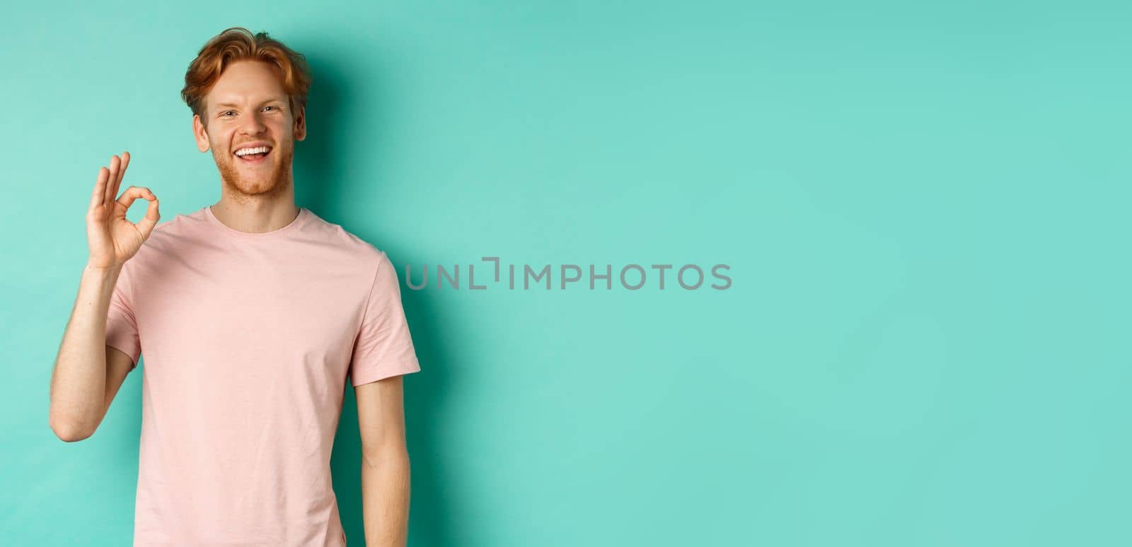 Handsome young bearded man in t-shirt showing Ok sign, smiling with white teeth and saying yes, agree with you, standing over turquoise background.