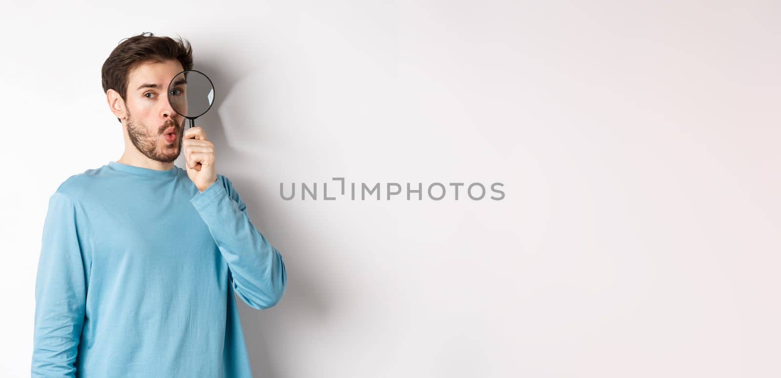 Surprised man found something, looking through magnifying glass with amazed face, standing over white background.