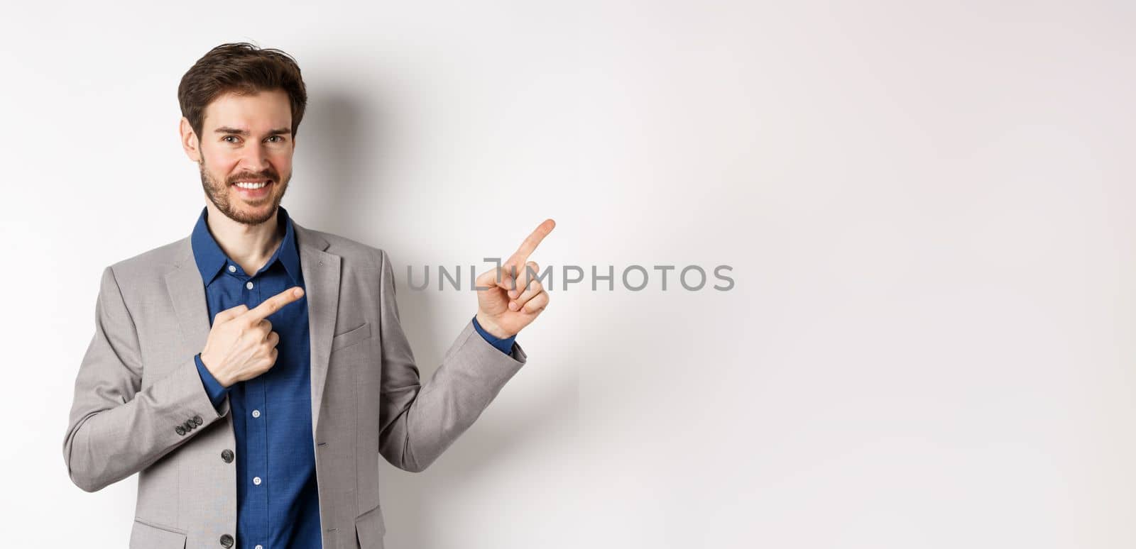 Handsome smiling man in business suit pointing fingers at upper right corner, showing logo, advertising company, standing on white background by Benzoix