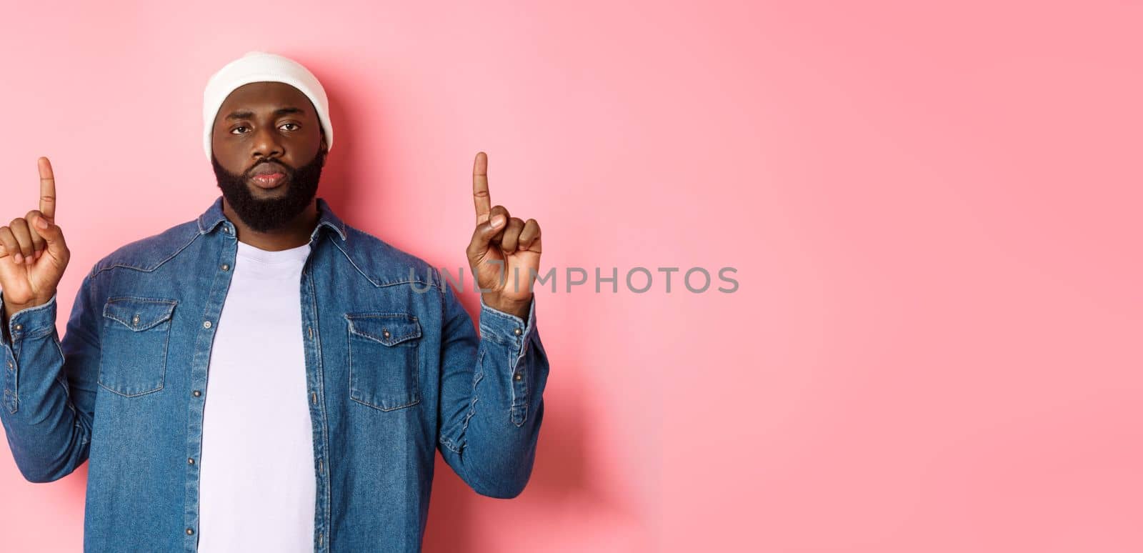 Serious and bothered Black man with beard, staring at camera and pointing fingers up, showing promo, standing over pink background by Benzoix