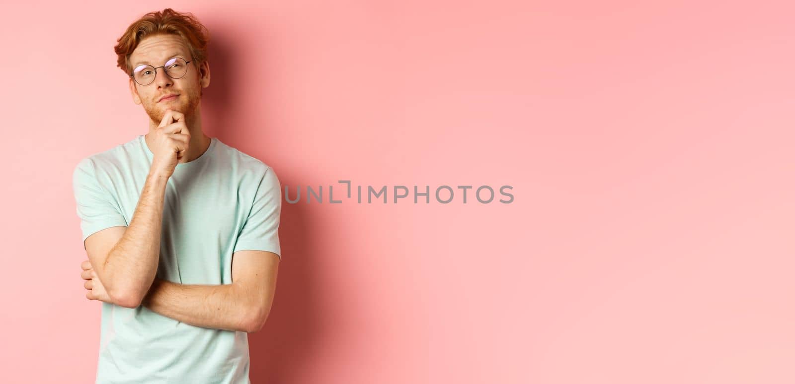Pensive handsome redhead man in glasses looking at upper right corner, touching chin and thinking, standing over pink background.