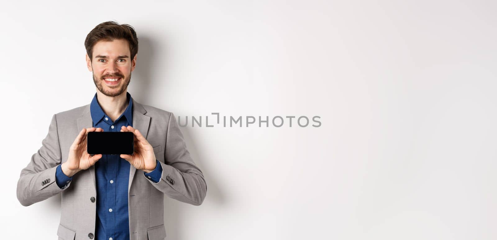 E-commerce and online shopping concept. Happy guy in business suit showing empty smartphone screen horizontally, smiling pleased at camera, white background by Benzoix