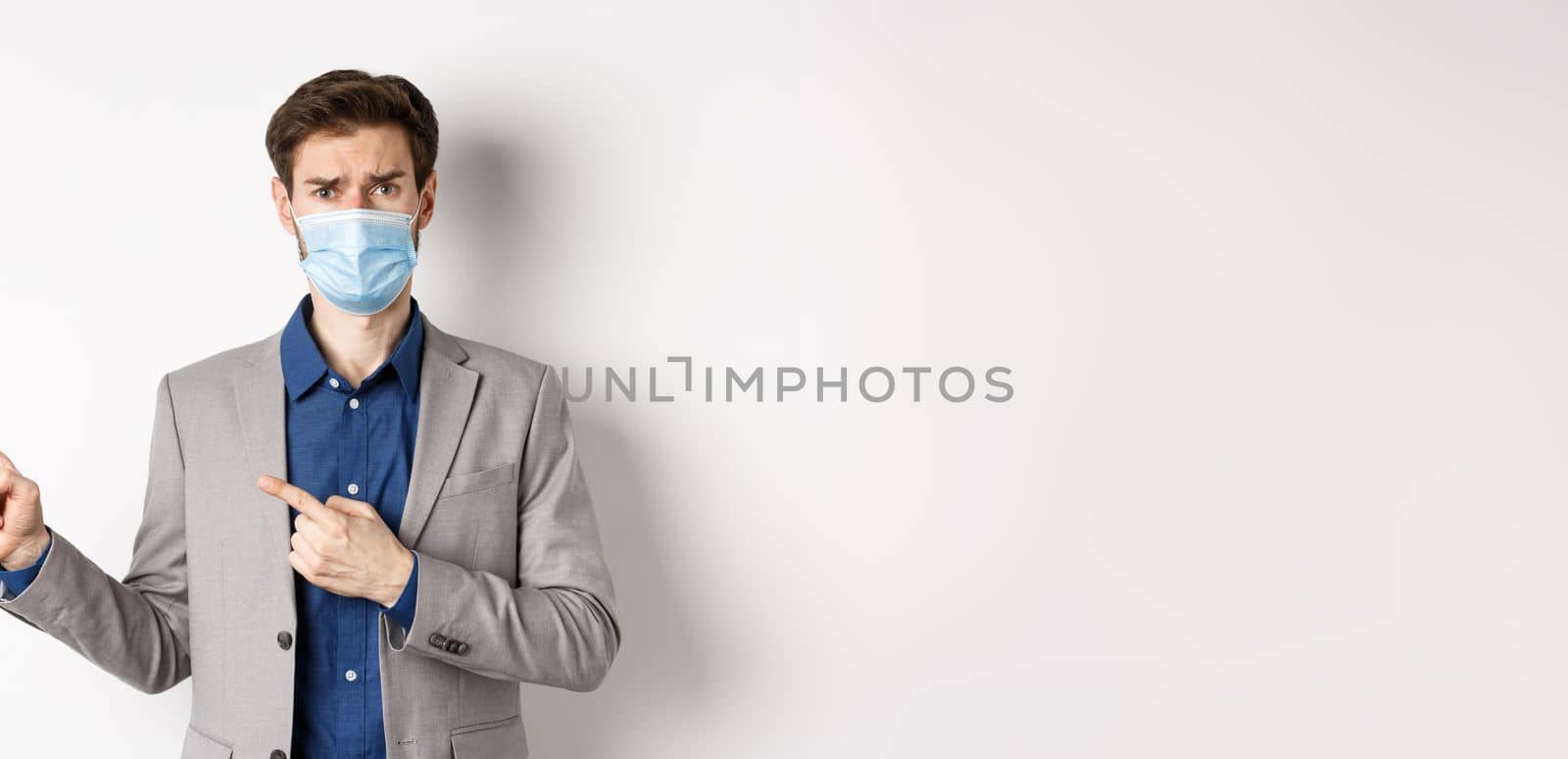 Covid-19, pandemic and business concept. Upset man in medical mask and office suit, frowning sad and pointing left at banner, standing on white background by Benzoix