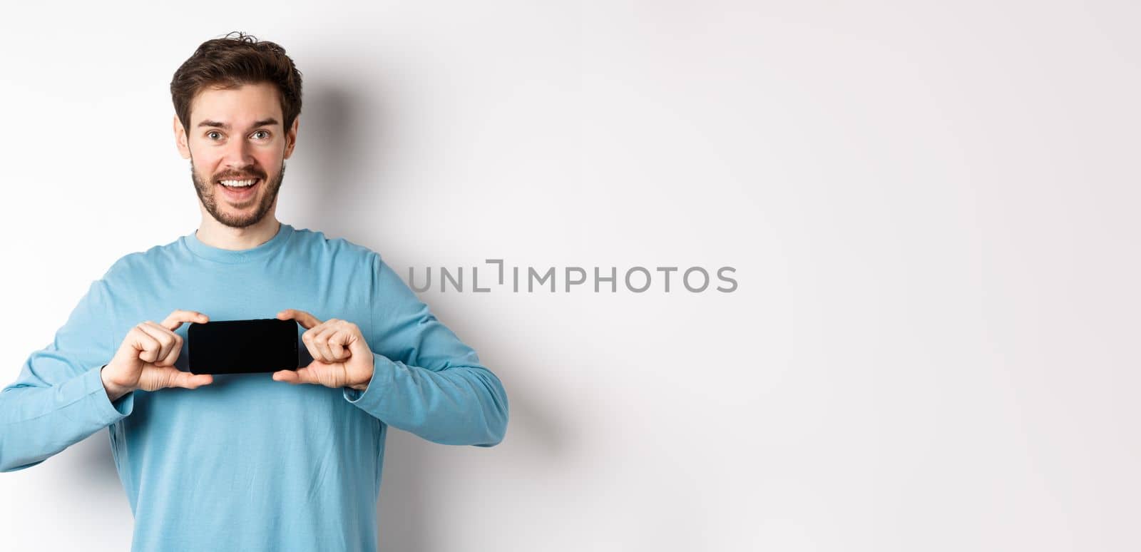 E-commerce and shopping concept. Smiling cheerful man showing empty smartphone screen in horizontal position, looking happy at camera, white background by Benzoix