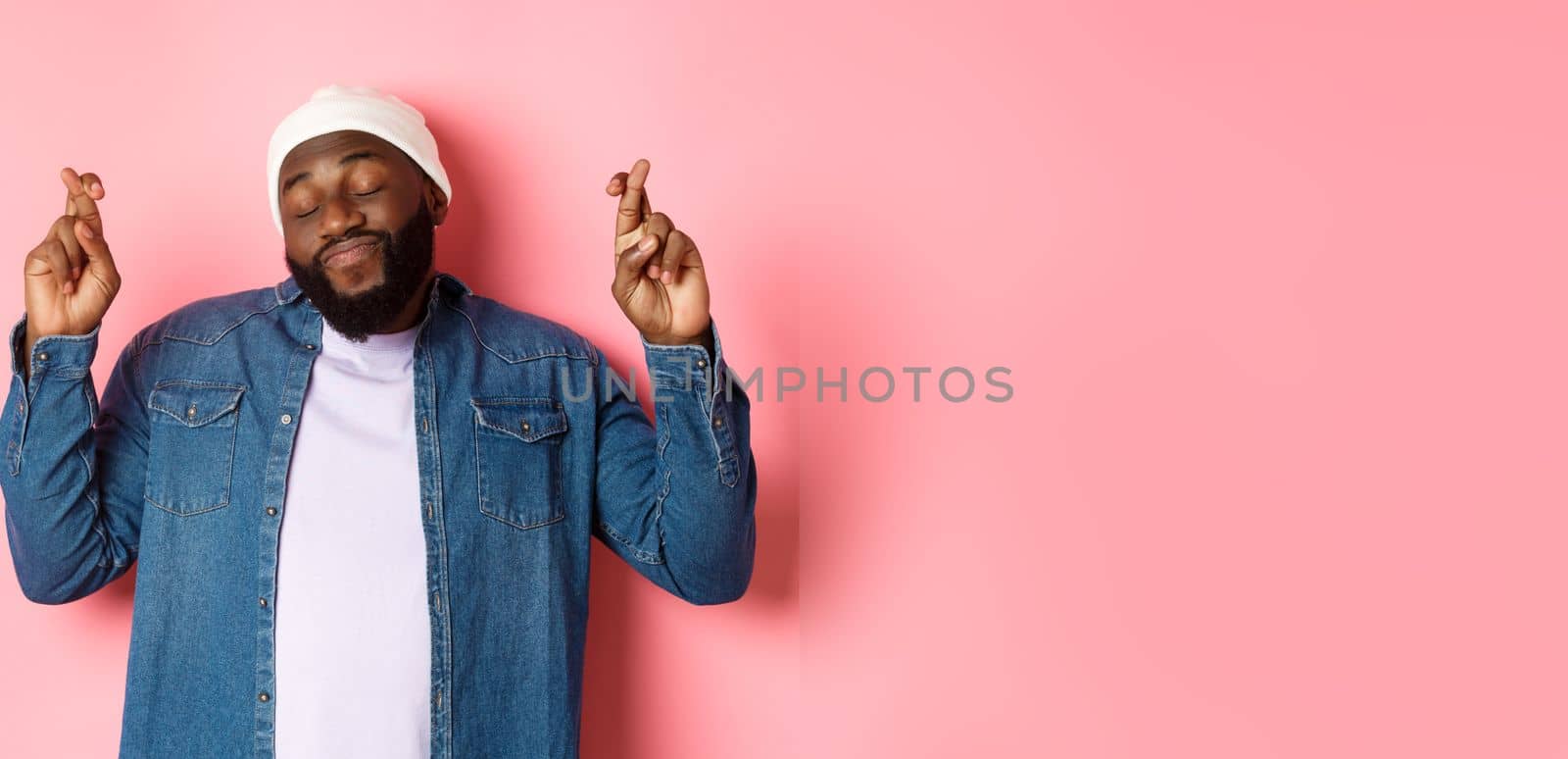 Hopeful and dreamy Black man making wish, cross fingers and smiling, standing over pink background by Benzoix