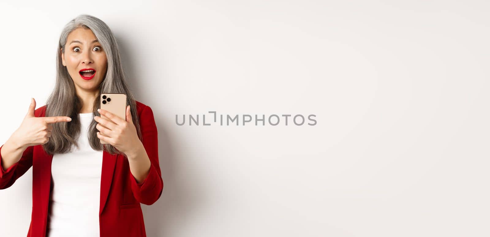Amazed asian lady in red blazer pointing finger at smartphone, staring impressed at camera, showing something online, white background.