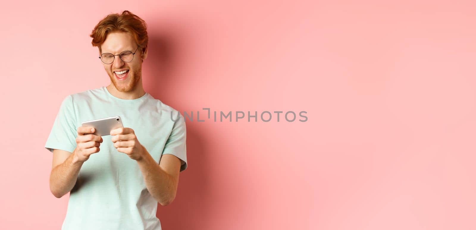 Happy young man with red messy haircut, wearing glasses, playing video game on smartphone and having fun, looking at mobile screen, standing over pink background.