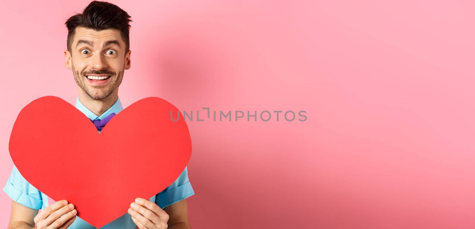 Hopeful man in love showing red heart sign, smiling at camera, waiting for soulmate on Valentines day, standing on pink background by Benzoix