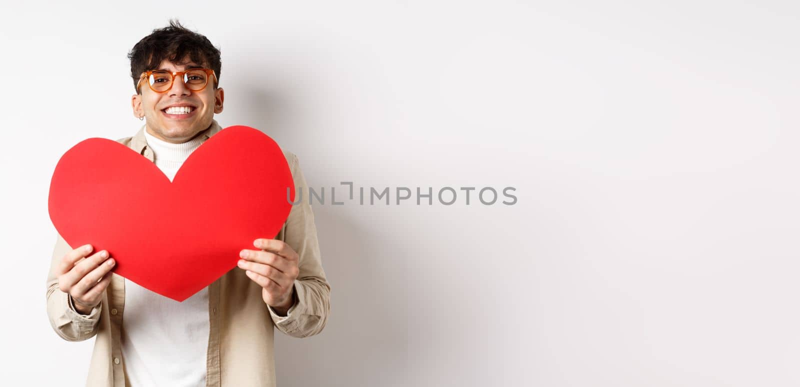Cheerful young man smiling and looking thrilled at camera, showing big red heart cutout on Valentines day, making romantic gift to lover, white background by Benzoix