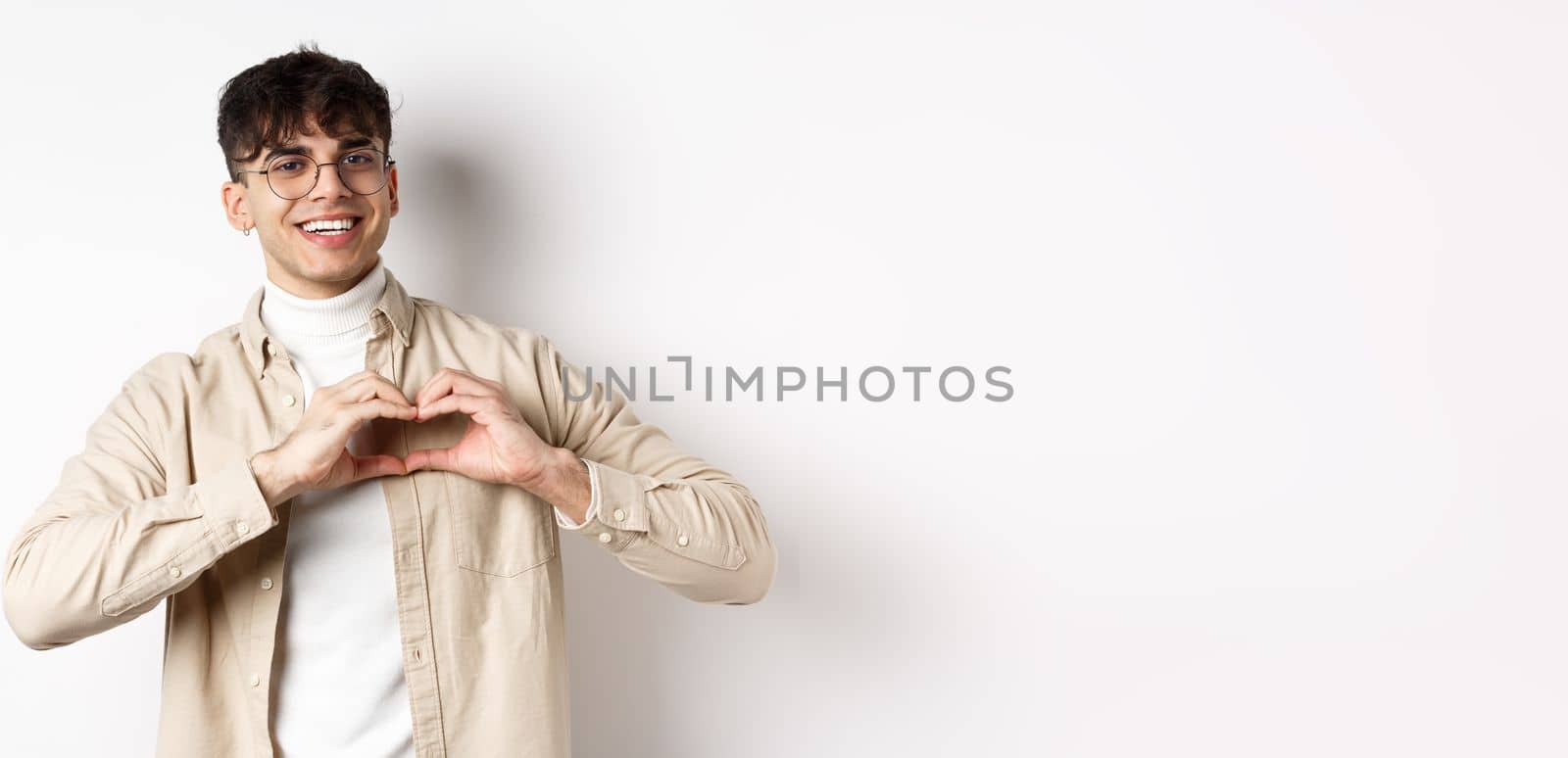 Happy Valentines. Handsome young man in glasses say I love you, showing heart sign and smiling at girlfriend, celebrating lovers day, standing over white background by Benzoix