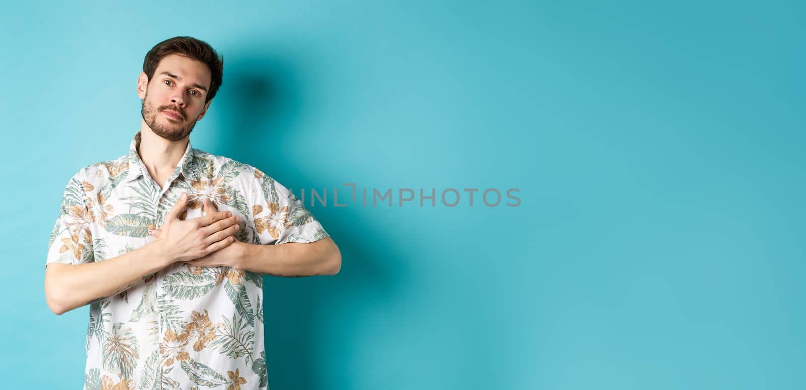 Handsome young man in summer shirt, holding hands on heart and looking thankful, being grateful, standing on blue background.