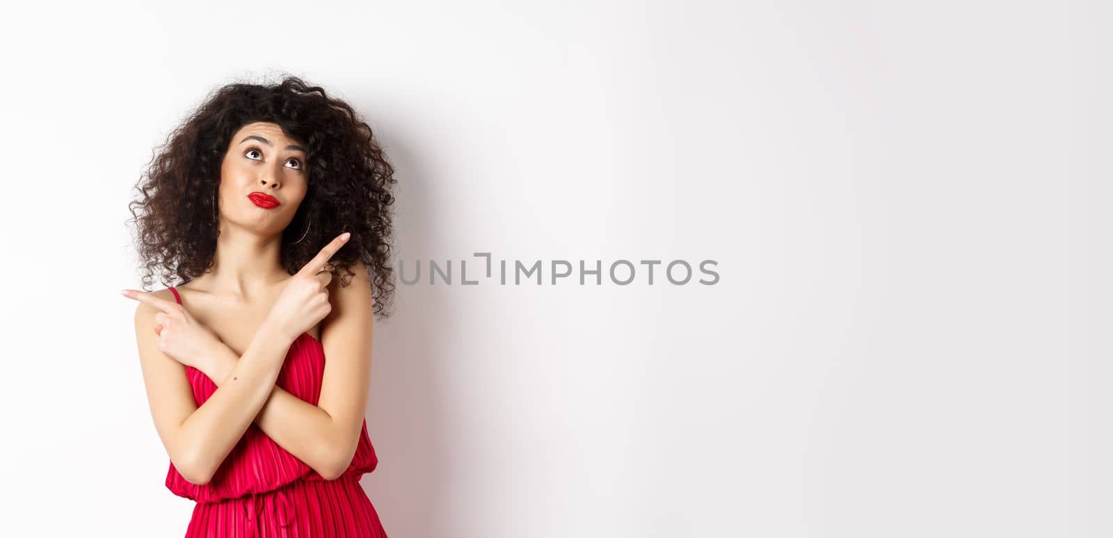 Indecisive silly woman in red dress, cross fingers and pointing sideways, looking up pensive, making choice, standing over white background by Benzoix