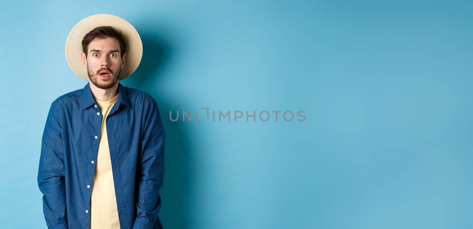 Young man tourist in summer hat drop jaw, staring at something shocking, standing on blue background by Benzoix