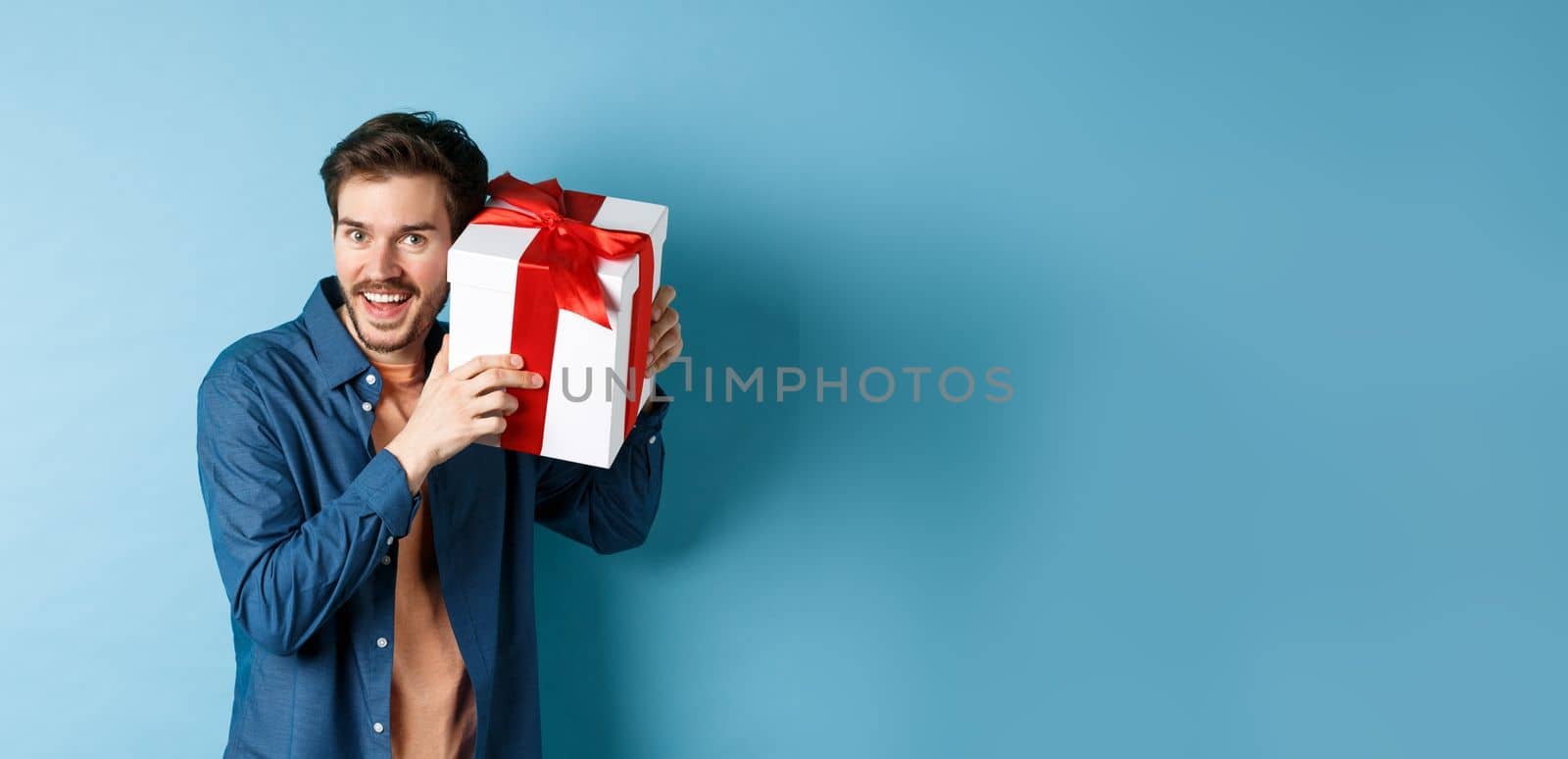 Valentines day. Happy young man got present on special holiday, trying guess what inside gift box and smiling, standing over blue background by Benzoix