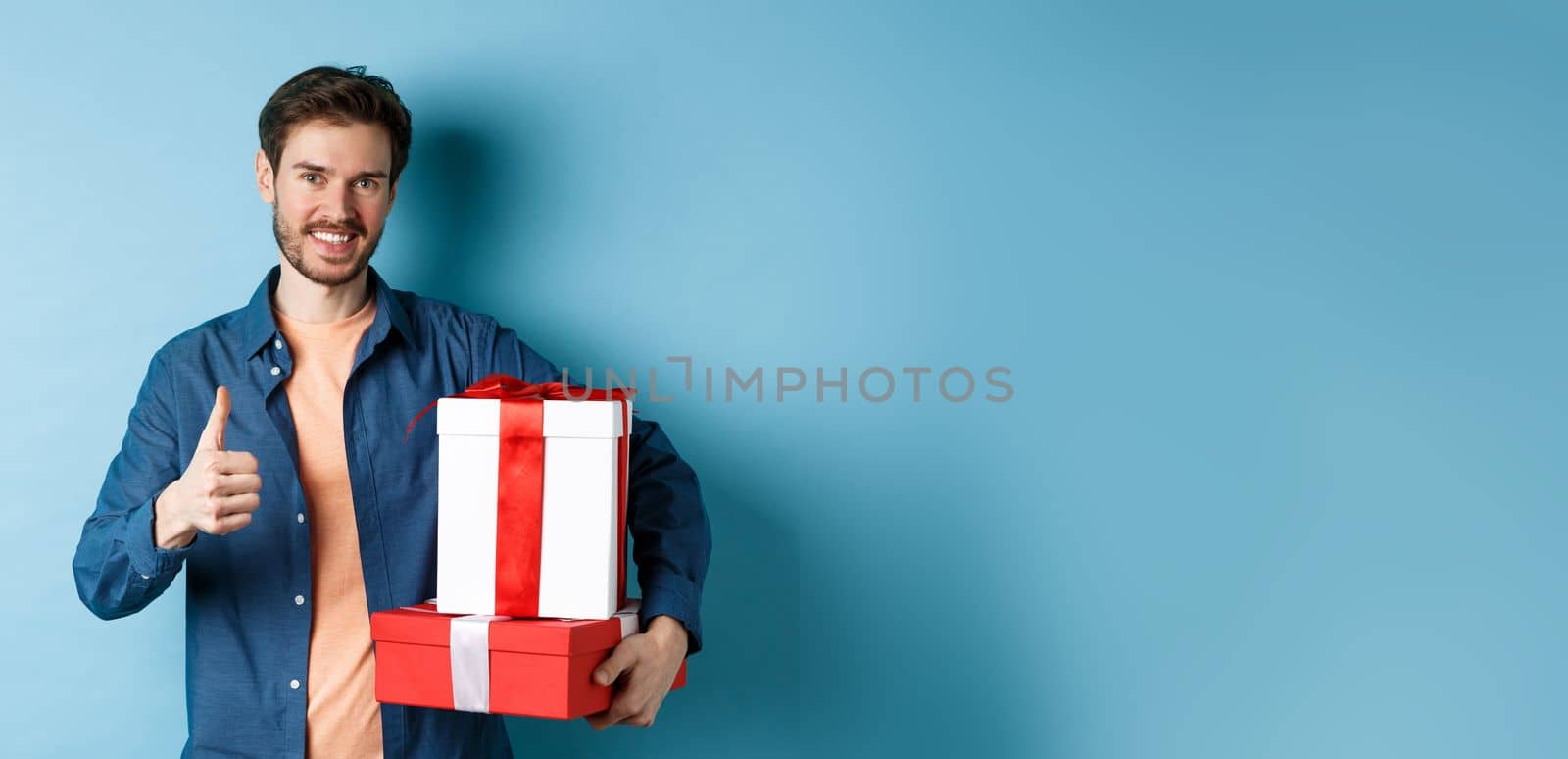 Smiling man holding romantic gifts and showing thumbs-up, celebrating Valentines day, buying presents for lover, standing over blue background by Benzoix