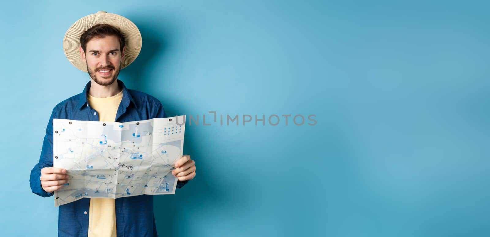 Happy and positive guy on vacation, looking at camera and holding map, smiling excited, going on summer travel, standing on blue background.