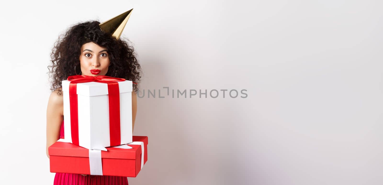 Cute birthday girl with curly hair and party hat, holding gifts and looking happy at camera, standing against white background.