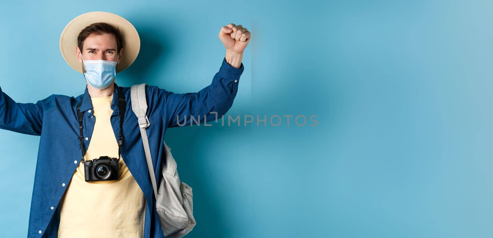 Covid-19 and summer holidays concept. Happy smiling tourist celebrating vacation, wearing face mask, shouting yes with joy and raising hands up, standing on blue background.