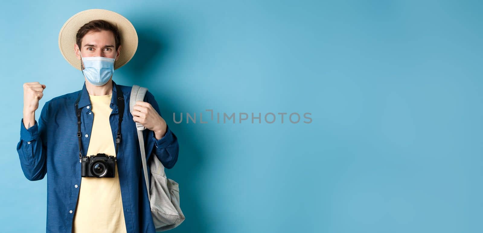 Covid-19 and summer holidays concept. Happy young man travelling abroad in medical mask, saying yes and raising fist up joyfully, cheerful about vacation, blue background by Benzoix