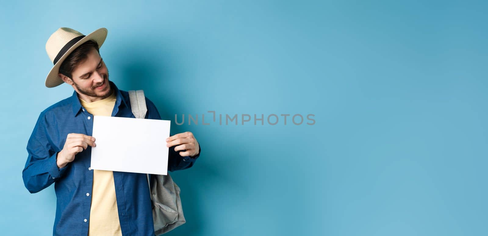 Happy tourist in straw hat smiling and looking at piece of paper for your logo, standing on blue background.