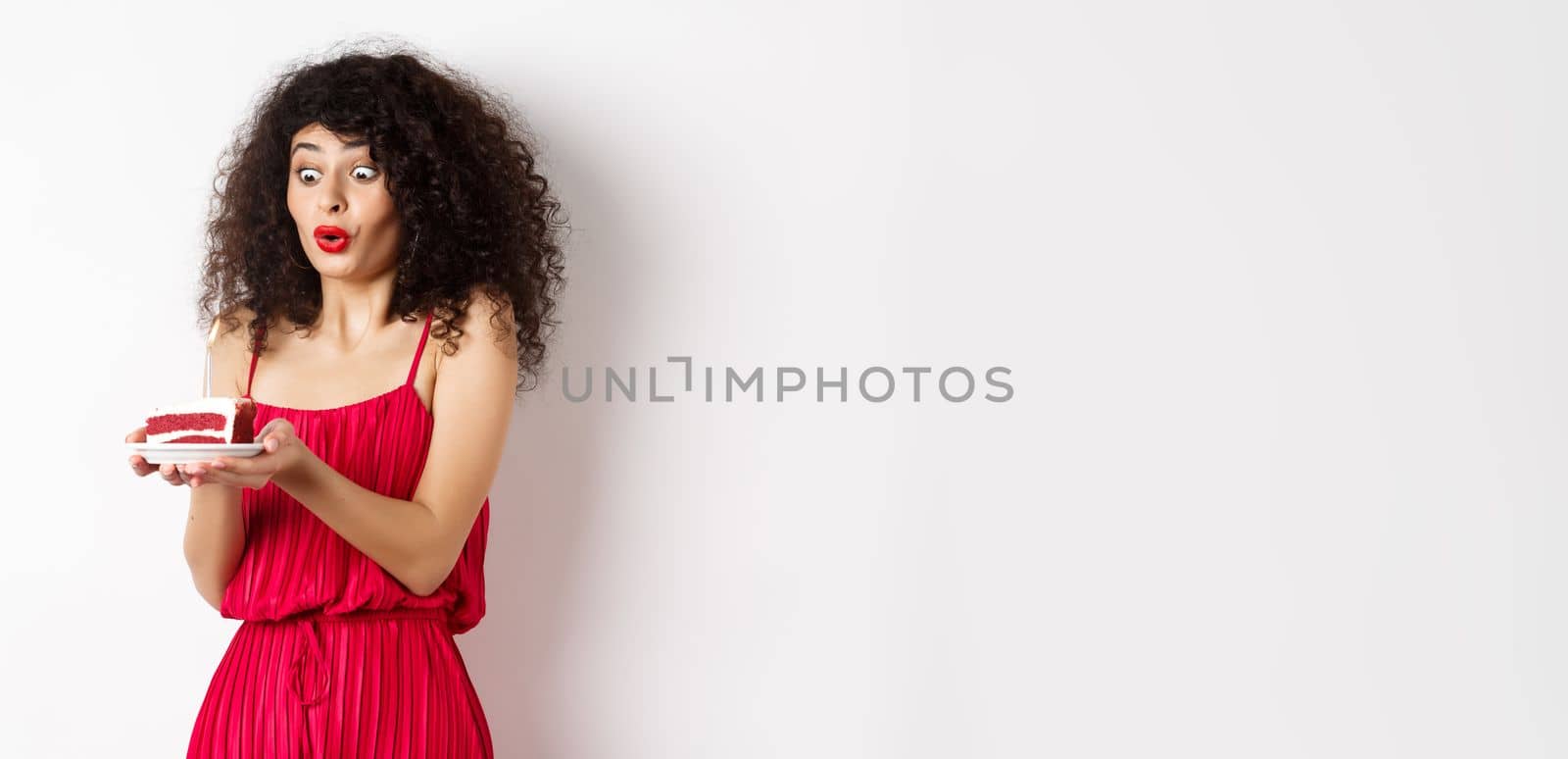 Excited birthday girl in red dress blowing candle on cake and making wish, standing on white background.