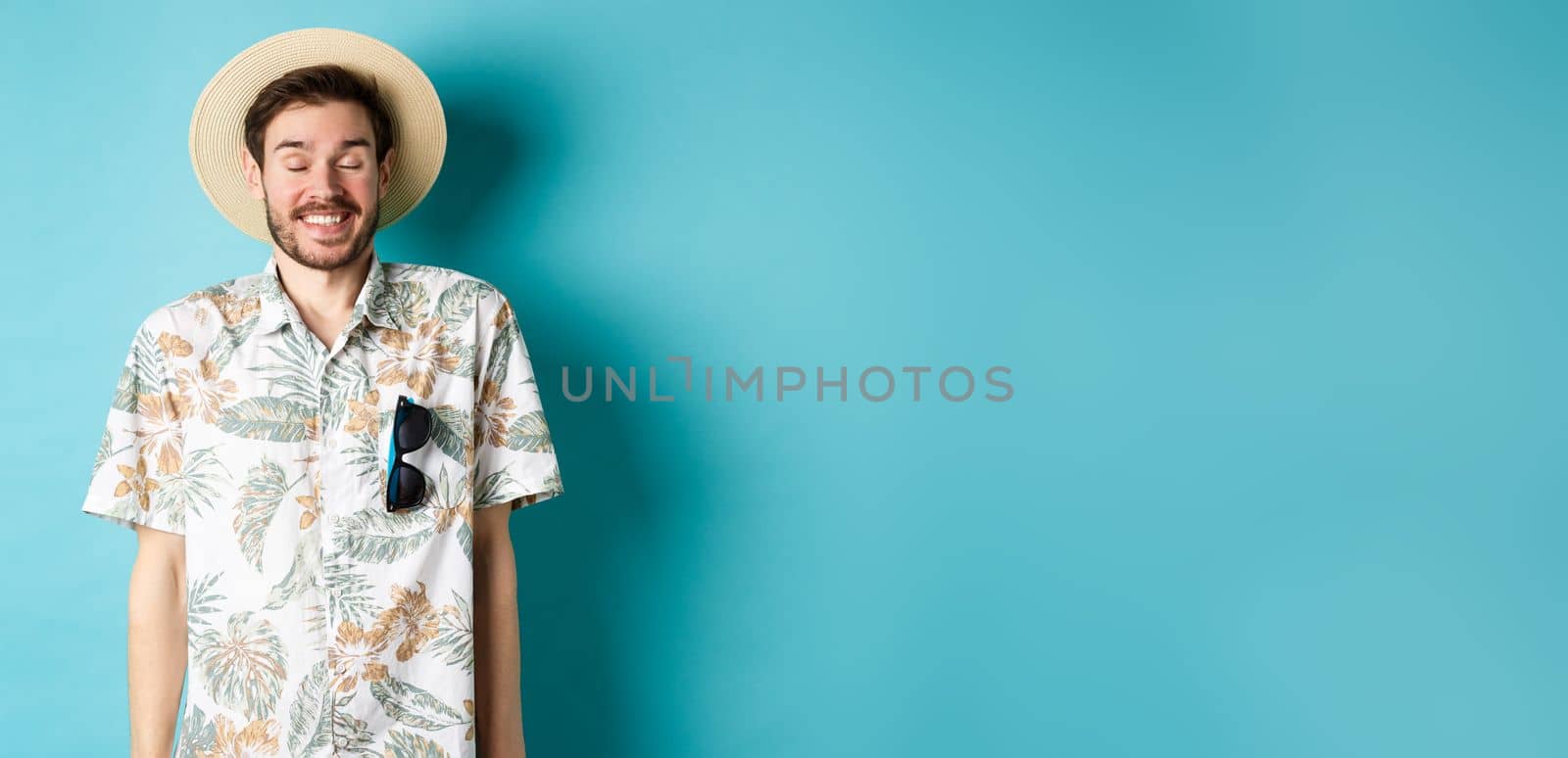 Happy tourist enjoying summer holiday, wearing straw hat and hawaiian shirt, standing joyful on blue background.