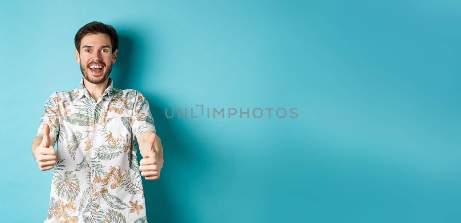 Excited smiling guy in hawaiian shirt show thumbs up, looking amazed, checking out cool promo, standing on blue background.