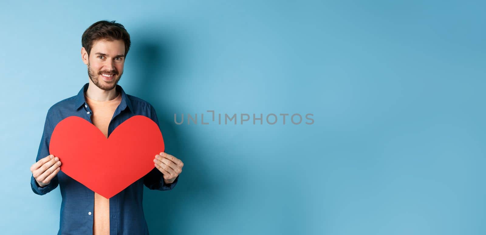 Handsome young man smiling, showing big red heart postcard for Valentines day, looking at camera happy, standing against blue background.