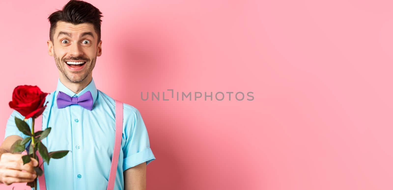 Valentines day and romance concept. Happy smiling man giving you red rose on romantic date, standing on pink background in bow-tie and shirt by Benzoix