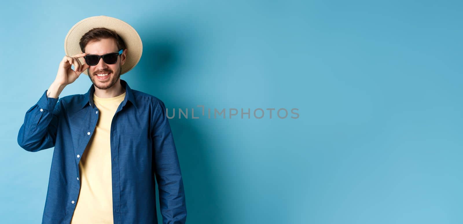 Happy smiling guy going on summer vacation, wearing straw hat and black sunglasses, standing on blue background.