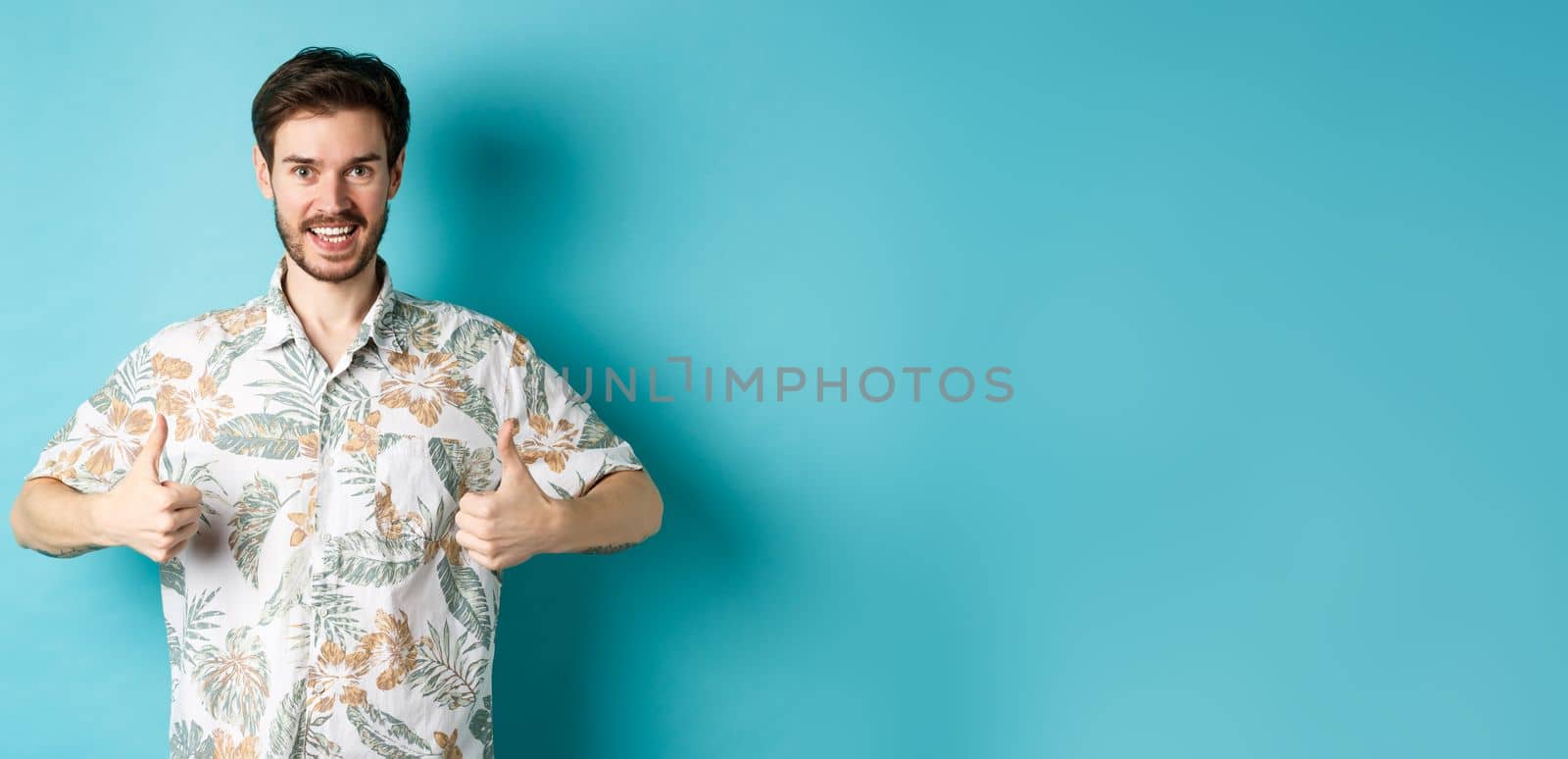 Handsome tourist showing thumbs up and say yes, praising good tourism agency, wearing hawaiian shirt, standing on blue background by Benzoix