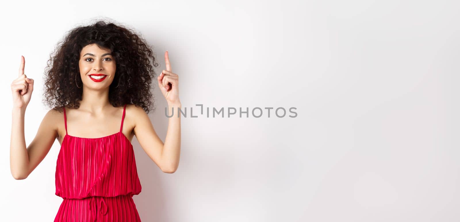 Smiling woman in elegant red dress and makeup, pointing fingers up and showing promo offer on valentines day, standing over white background.