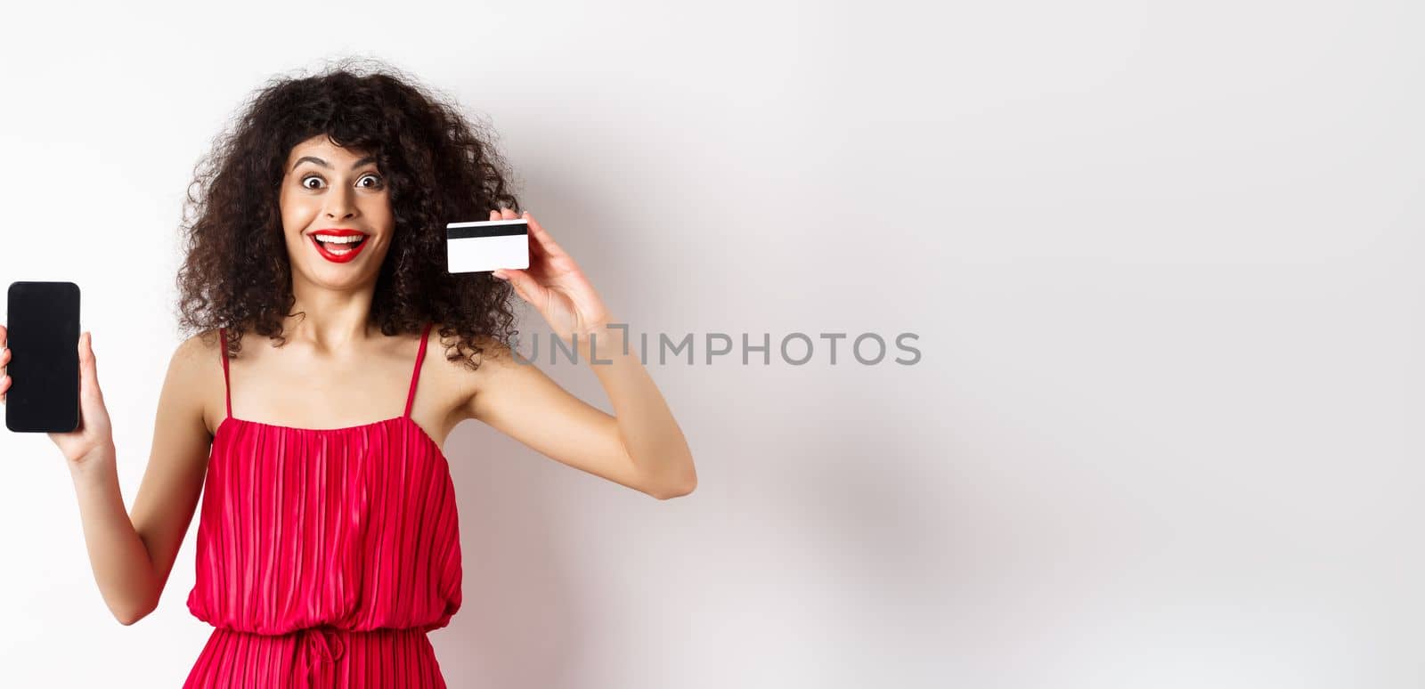 Online shopping concept. Excited curly-haired woman in red dress showing empty smartphone screen and plastic credit card, smiling happy at camera, standing on white background by Benzoix