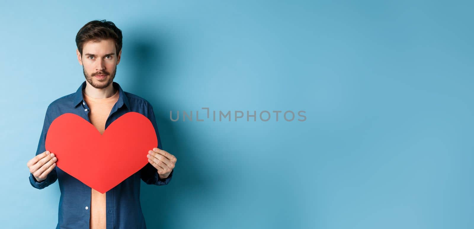 Young man looking for soulmate on valentines day, holding big red heart and looking at camera, standing over blue background.