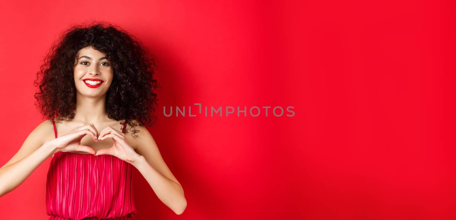Valentines day. Romantic girl with curly hairstyle in evening dress, smiling and showing heart sign, say I love you on lovers holiday, standing over red background.