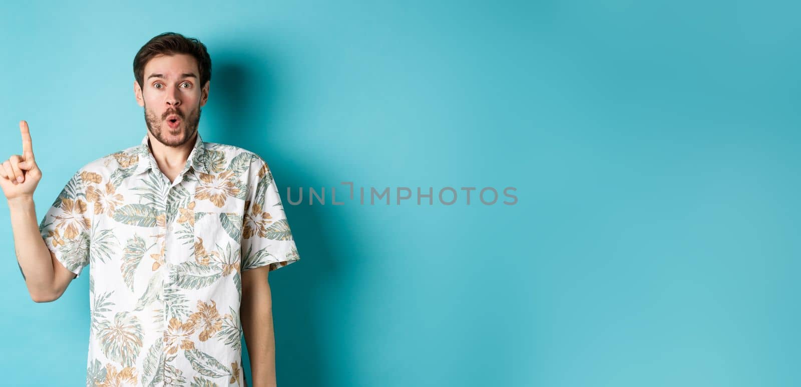 Surprised caucasian tourist in hawaiian shirt, pointing finger up and say wow, checking out promotion, standing on blue background by Benzoix