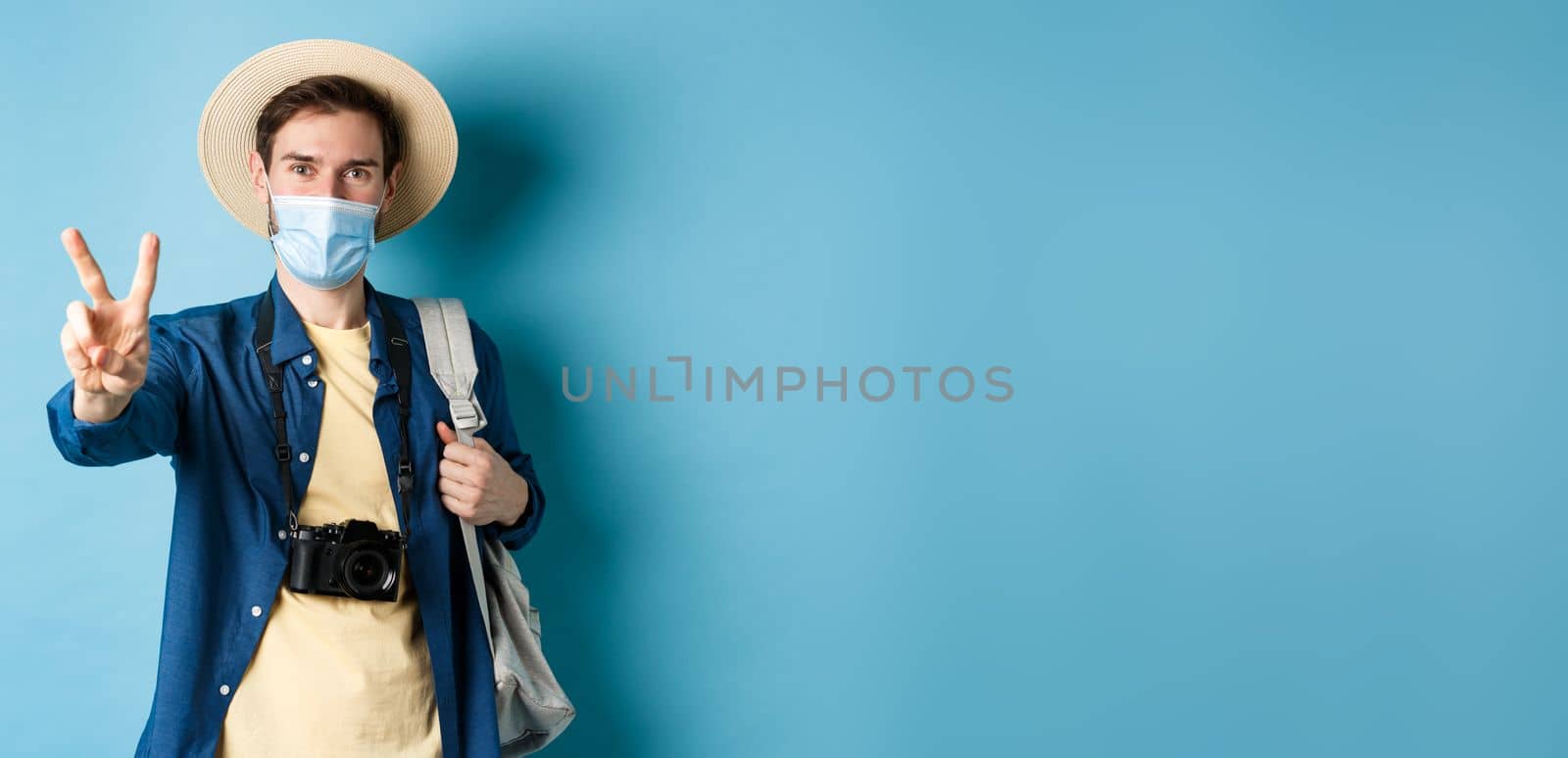 Covid-19, pandemic and travel concept. Positive guy going on vacation, wearing summer hat and medical mask, showing peace sign, standing with backpack and camera on blue background by Benzoix