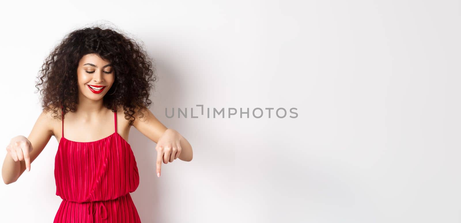 Cheerful elegant woman in red dress and makeup, looking and pointing down with pleased smile, showing advertisement, standing over white background.