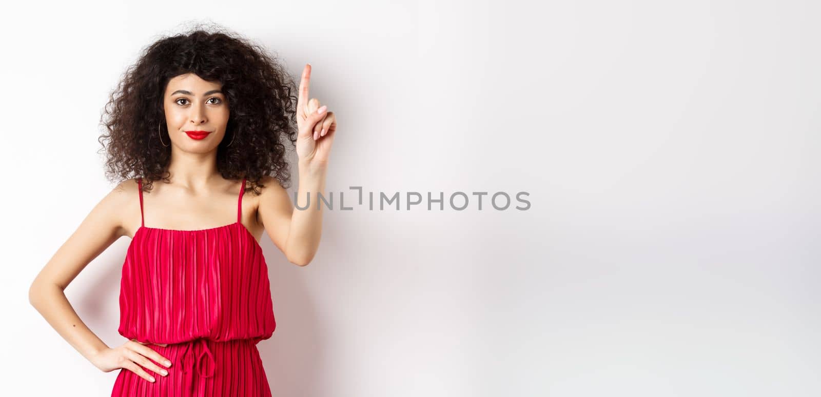 Smiling caucasian woman with curly hair, wearing red dress, showing rule number one gesture, raising finger and looking confident at camera, white background.