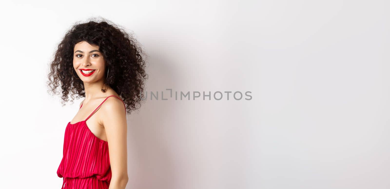 Profile of fashionable curly-haired woman in red dress, turn head at camera and smiling confident, standing over white background.