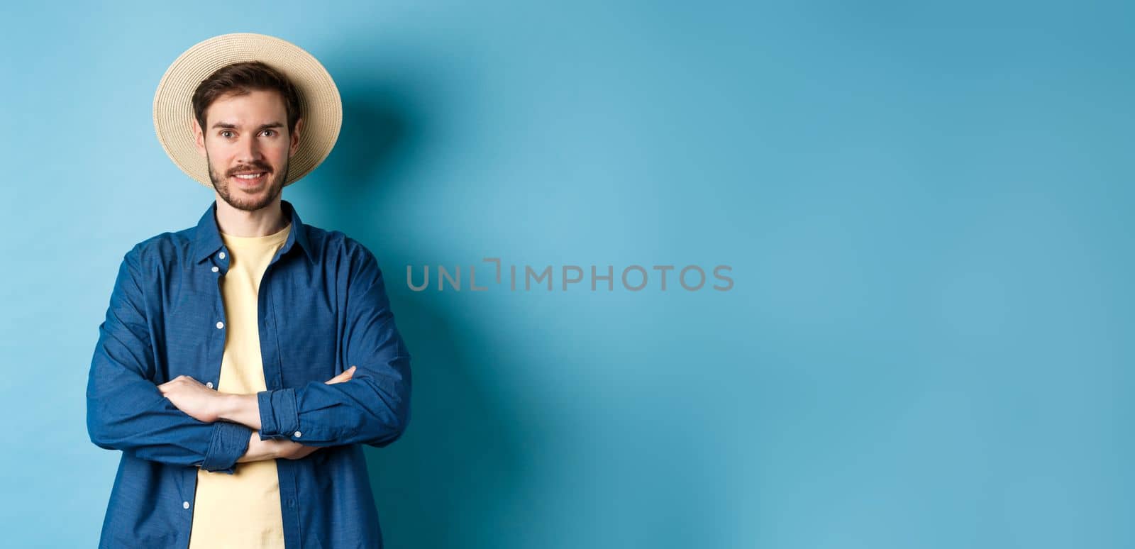 Portrait of cheerful guy going on vacation in straw summer hat, cross arms on chest and smiling, looking confident, standing on blue background by Benzoix