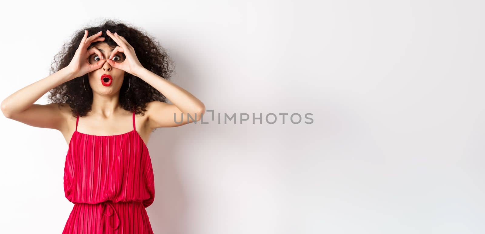 Impressed young woman seeing something awesome, look through hand binoculars and say wow, standing amazed at white background.