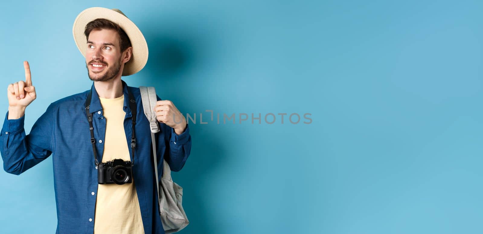 Handsome positive guy on vacation, wearing summer hat, looking and pointing up at logo, smiling pleased, standing with camera and backpack on blue background.