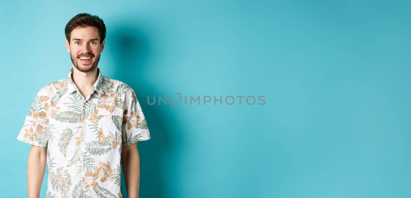 Cheerful caucasian guy going on vacation, wearing summer shirt and smiling, standing on blue background.