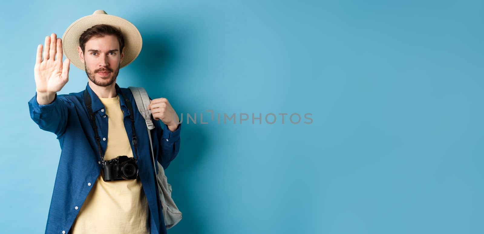 Serious male tourist warn you, stretch out hand to show stop gesture. Man on vacation forbid something, refusing or prohibiting action, standing with backpack on blue background.